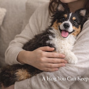 May include: A tricolor puppy with black, white, and tan fur is held in someone's arms. The puppy has a pink tongue and is smiling. The text at the bottom of the image reads, "A Hug You Can Keep Forever."