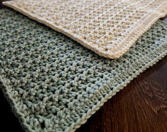 May include: Two handmade crochet washcloths. One is a light sage green color, and the other is a cream color. Both are square and have a textured stitch pattern. The washcloths are displayed on a dark brown wooden surface.