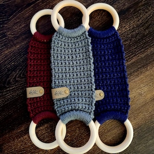 May include: Three crocheted teethers with wooden rings. The teethers are in burgundy, gray, and navy blue. Each teether has a small leather tag with a leaf design.