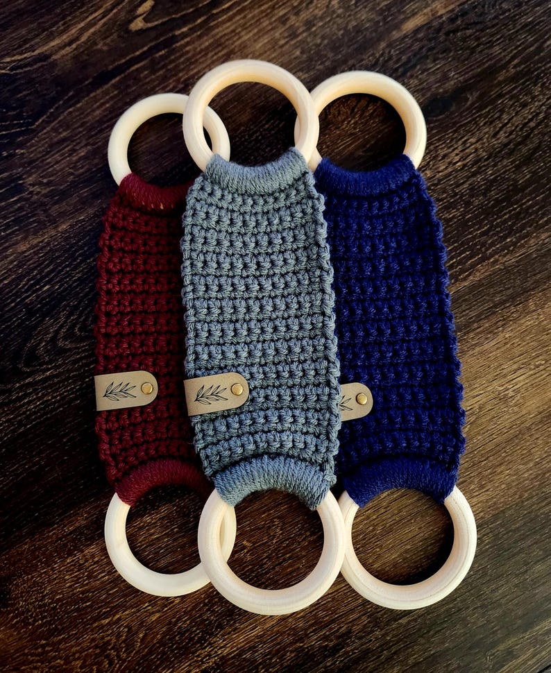 May include: Three crocheted teethers with wooden rings. The teethers are in burgundy, gray, and navy blue. Each teether has a small leather tag with a leaf design.