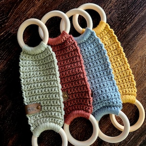 May include: Four crocheted teethers with wooden rings. The teethers are in shades of green, red, blue, and yellow. Each teether has a leather tag with a small leaf design.
