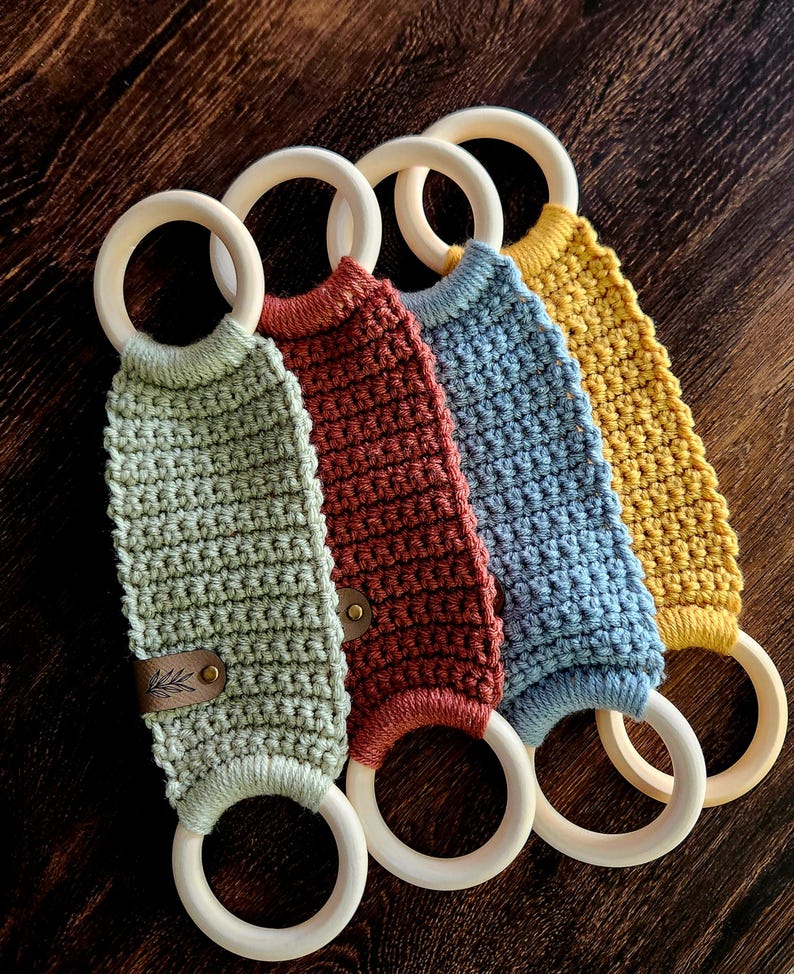 May include: Four crocheted teethers with wooden rings. The teethers are in shades of green, red, blue, and yellow. Each teether has a leather tag with a small leaf design.