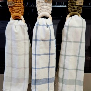 May include: Three kitchen towels hanging on a rack. The towels are white with a plaid pattern in blue, green, and brown. Each towel is attached to a wooden ring with a crocheted loop in a different color: mustard yellow, cream, and olive green.