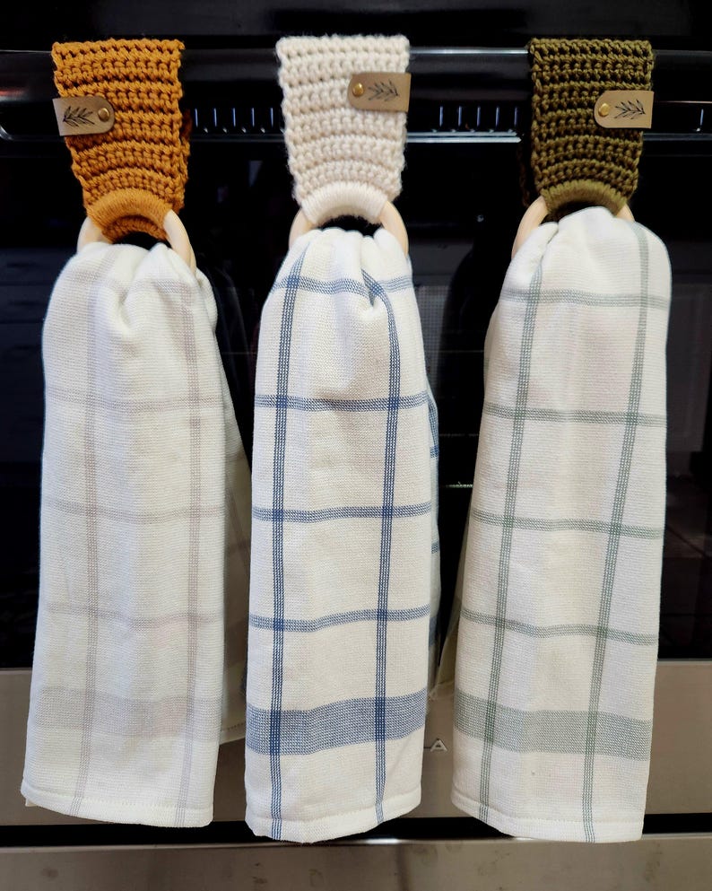 May include: Three kitchen towels hanging on a rack. The towels are white with a plaid pattern in blue, green, and brown. Each towel is attached to a wooden ring with a crocheted loop in a different color: mustard yellow, cream, and olive green.