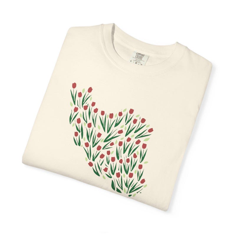May include: Cream-colored long-sleeve shirt featuring a floral design of red tulips with green stems and leaves. The tulip arrangement is on the left side of the shirt. The shirt is folded and has a tag.