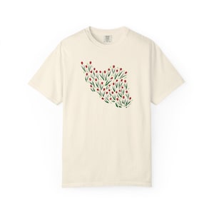 May include: Cream-colored t-shirt featuring a floral design in the shape of a country. The design is composed of red flowers with green stems and leaves. The shirt has short sleeves and a crew neck.
