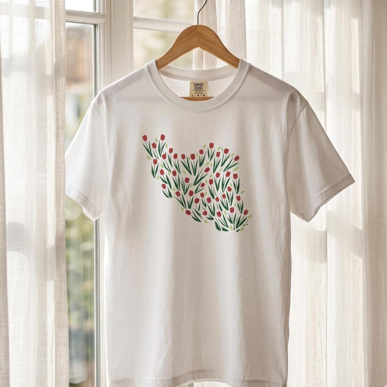 May include: White t-shirt with a crew neck, featuring a floral design of red flowers and green leaves. The design is arranged in a shape resembling a country. The shirt is hanging on a wooden hanger.