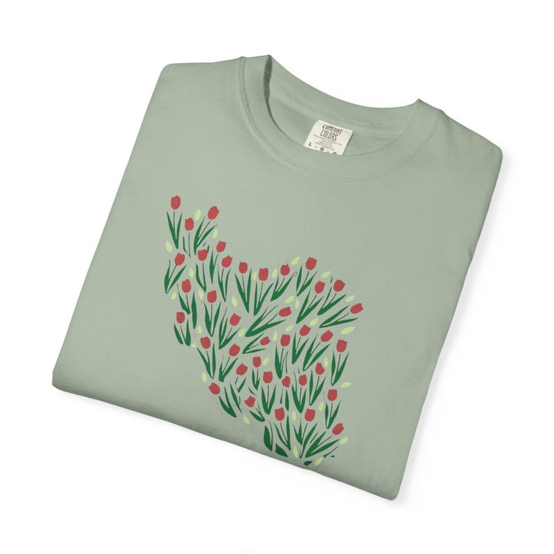 May include: Sage green t-shirt featuring a floral design of red tulips with green stems and leaves. The graphic is arranged in a stylized shape. The shirt is folded and has a tag in the upper right corner.