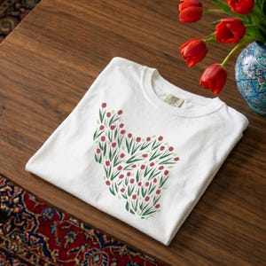 May include: White t-shirt with a floral design of red tulips and green leaves. The shirt is folded on a wooden table, with a vase of red tulips and a patterned rug in the background. The shirt has a Comfort Colors tag.