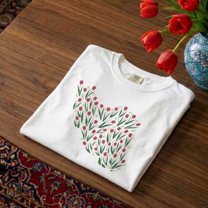 May include: White t-shirt with a floral design of red tulips and green leaves. The shirt is folded on a wooden table, with a vase of red tulips and a patterned rug in the background. The shirt has a Comfort Colors tag.