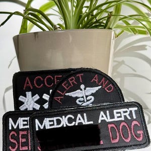 May include: Three rectangular black embroidered patches with white and pink stitching. One patch features a white caduceus symbol and the text ALERT. Another displays a white star of life. The third patch reads MEDICAL ALERT DOG in white and pink.
