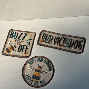 May include: Three embroidered patches on a light background. One square patch reads BUZZ OFF with a bee, one rectangular patch reads SERVICE DOG with floral accents, and one circular patch reads BUSY AND WORKING LEAVE US BEE with a bee.