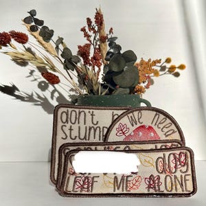 May include: Three rectangular and semi-circular embroidered patches with brown borders on a light fabric. The patches feature brown text reading "don't be stumped", "we need", and "LEAF ME ALONE" with red mushroom and autumn leaf illustrations.