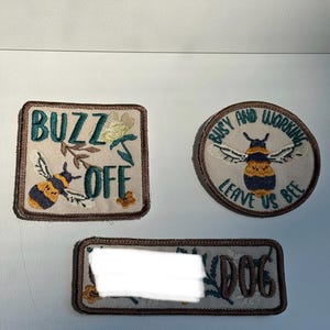 May include: Three embroidered fabric patches with brown borders. One square patch reads BUZZ OFF with a bee and floral design. One circular patch reads BUSY AND WORKING LEAVE US BEE with a bee. One rectangular patch features floral art and the word DOG.