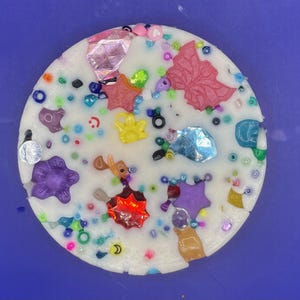 以下が含まれることがあります： カラフルな装飾が施された、丸く白い装飾品。星、蝶、花の模様など、さまざまな形があり、散らばったビーズや虹色のアクセントも特徴です。この作品は、無地の青色の背景に配置されています。