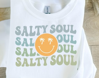 May include: White sweatshirt featuring the text "SALTY SOUL" in blue, green, and sage green. An orange smiley face with palm trees replaces the "O" in "SOUL". The Gildan brand tag is visible.