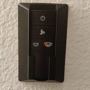 May include: A black wall-mounted remote control with five buttons. The top button has a power symbol. The second button has a fan symbol with three speed settings. The third button has a light symbol. The fourth button has a light symbol with a rainbow arc.