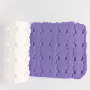 Può includere: Un mattarello cilindrico bianco con un motivo in rilievo di impronte di zampe. Accanto al rullo si trova un pezzo rettangolare piatto di argilla viola impresso con lo stesso motivo, che mostra la texture creata dallo strumento.
