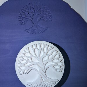 Può includere: Un timbro circolare bianco con un design ad albero in rilievo posizionato su una superficie viola piatta. Il timbro ha creato un'impronta profonda e incavata dello stesso motivo ad albero direttamente nel materiale viola, mostrando il rilievo.