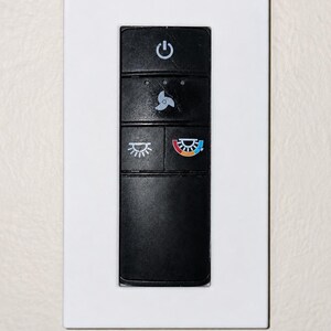 May include: A rectangular wall-mounted control panel featuring a white outer frame and a black central interface. The black section includes a power button, a fan speed icon, a light brightness icon, and a colour temperature adjustment icon.
