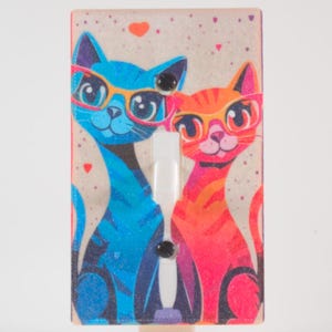 Può includere: Una copertura per interruttore della luce con un'illustrazione di gatto blu e arancione con occhiali. I gatti sono ambientati su uno sfondo maculato con accenti a cuore. L'interruttore è bianco.