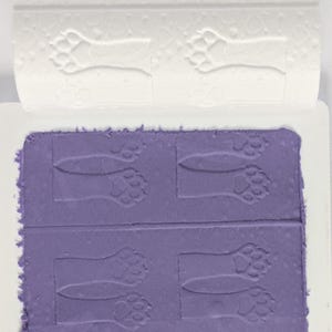 Peut inclure: Un rouleau à pâtisserie cylindrique blanc avec un motif en relief de pattes d'animaux, placé au-dessus d'une plaque rectangulaire violette présentant le même motif gaufré. L'outil sert à créer des empreintes répétitives sur des surfaces.