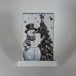 May include: A decorative light switch cover featuring a black and white illustration of a snowman wearing a top hat and scarf, standing next to a Christmas tree. The design includes a star and a bird. The cover is mounted on a white base.
