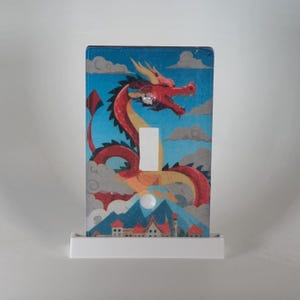 Könnte beinhalten: Dekorative Lichtschalterabdeckung mit einem rot-gelben Drachen vor blauem Himmel mit Wolken. Das Drachendesign umfasst eine Bergkette und eine kleine Stadt am unteren Rand. Der Schalter ist weiß.