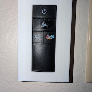 Puede incluir: Una placa de pared rectangular blanca con un panel de control negro. El panel de control tiene cuatro botones con iconos para encendido, ventilador, luz y temperatura. La placa está montada en una pared clara.