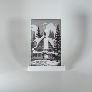 May include: A decorative light switch cover featuring a winter scene with a snow-covered house, pine trees, and a starry sky. The switch is in the center of the cover, with a white base. The artwork is in shades of gray and white.