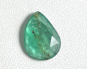 May include: A pear-shaped, faceted emerald gemstone with a vibrant green hue. The gemstone has a polished surface and a triangular facet near the bottom. The stone is set against a white textured background.