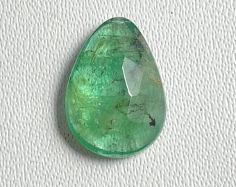 May include: A pear-shaped, faceted emerald gemstone. The gem is a vibrant green color with internal inclusions and a polished surface. The stone is set against a white textured background.