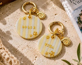 May include: Two round keychains featuring yellow and white vertical stripes. One keychain displays the gold text DOLCE, the other VITA. Both include gold-toned metal rings and small gold-colored seashell and starfish charms attached to the circular discs.