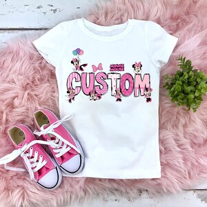 May include: White t-shirt with a pink and black "CUSTOM" design featuring Minnie Mouse characters and the words "MINNIE MOUSE". Pink canvas trainers with white laces are next to the shirt on a pink fluffy surface.