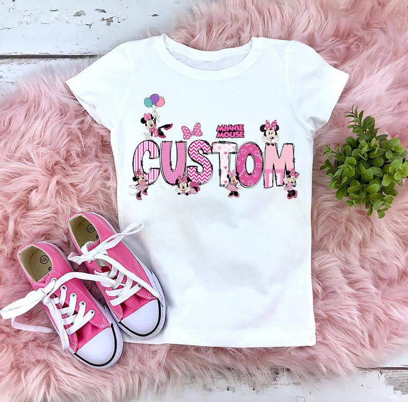 May include: White t-shirt with a pink and black "CUSTOM" design featuring Minnie Mouse characters and the words "MINNIE MOUSE". Pink canvas trainers with white laces are next to the shirt on a pink fluffy surface.