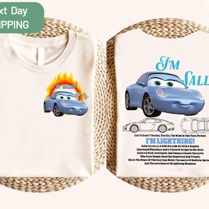 May include: Cream-coloured t-shirt with a cartoon image of a light blue car and the text "I'm Sally". The front of the shirt has a car graphic with flames and the text "I'm Sally". The back of the shirt has a car graphic and text.