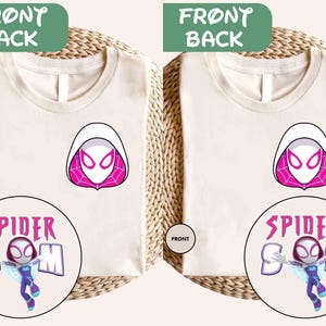 Puede incluir: Camisetas blanquecinas con un diseño de Spider-Gwen. Las camisetas muestran una máscara con capucha blanca con detalles en rosa y las palabras "SPIDER MOM" y "SPIDER BOSS" en un gráfico circular. Las camisetas se exhiben sobre una superficie tejida.