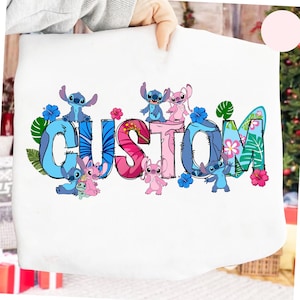 Pode incluir: Saco de mão branco com a palavra "CUSTOM" em letras coloridas, decorado com personagens de desenhos animados e flores tropicais. As letras são azuis, rosas e verdes, com personagens azuis e rosas. O design inclui folhas e flores tropicais.