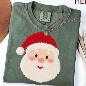 May include: A sage green t-shirt with a Santa Claus face design. The Santa has a red hat, white beard, and rosy cheeks. A silver necklace with a small pendant rests on the shirt. The shirt is made of a soft material and is perfect for the holiday season.
