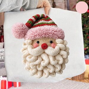 Peut inclure: Un coussin fait à la main représentant le Père Noël, avec un bonnet à pompon rose, un bonnet rayé rouge et vert, et une barbe crème. Le visage du Père Noël a un nez rouge, des joues roses et des yeux noirs. Le coussin est tenu par une personne.