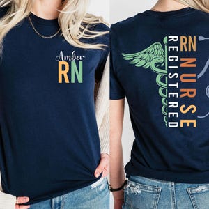 May include: A navy blue t-shirt with the text "Amber RN" on the front in white, orange, and green. The back displays a caduceus design and the words "RN Registered Nurse" with a stethoscope graphic.