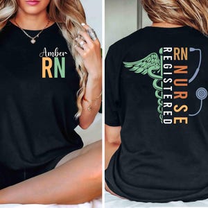 May include: Black t-shirt with "Amber RN" in orange and green on the front. The back features a green caduceus with wings, a stethoscope, and the words "RN NURSE REGISTERED" in orange, green, and white.