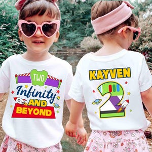 May include: Two white short-sleeved t-shirts featuring colorful graphic prints. One shirt displays the text "TWO Infinity AND BEYOND" with space-themed icons. The other shirt shows the name "KAYVEN" above a large number "2" with space motifs.