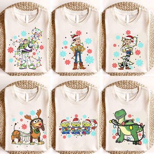 Könnte beinhalten: Sechs cremefarbene T-Shirts mit weihnachtlichen Toy Story-Charakteren. Buzz Lightyear, Woody, Jessie, Slinky Dog, die Aliens und Rex sind mit festlichen Lichtern und Schneeflocken dargestellt. Jedes Shirt liegt auf einer gewebten Matte.