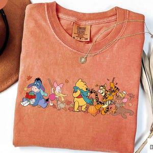 May include: Orange Comfort Colors t-shirt featuring a graphic of Winnie the Pooh and friends in autumn attire. The design includes Eeyore, Piglet, Winnie the Pooh, Tigger, and Roo. A gold necklace is draped over the shirt.