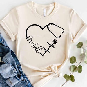 May include: A cream-coloured t-shirt with a black heart design incorporating a stethoscope and a heartbeat line. The name "Michelle" is written in cursive within the heart. The shirt has rolled-up sleeves and a relaxed fit.