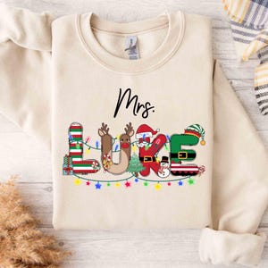 Puede incluir: Sudadera beige con la frase "Mrs. Luke" en un diseño festivo y colorido. Las letras están decoradas con elementos navideños como renos, Papá Noel y bastones de caramelo, con luces de cadena en la parte inferior.