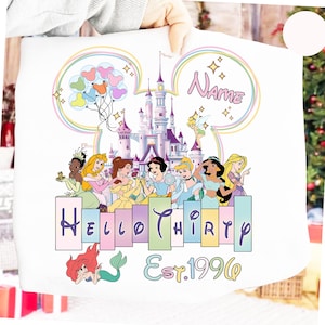 May include: White sweatshirt featuring a colorful graphic of Disney princesses, a castle, balloons, and the text "Hello Thirty Est. 1996". The design includes a space for a name, perfect for a birthday celebration.