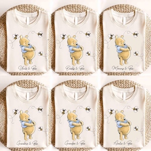 Puede incluir: Seis camisetas color crema con un diseño de oso de dibujos animados y el texto "Uncle to Bee", "Daddy to Bee", "Mommy to Bee", "Grandma to Bee", "Grandpa to Bee" y "Auntie to Bee". El oso lleva una camisa azul y está rodeado de abejas.
