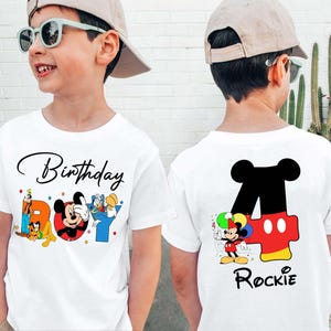 Könnte beinhalten: Weißes T-Shirt mit farbenfrohem Mickey Mouse-Geburtstagsdesign. Auf der Vorderseite stehen die Worte "Birthday Boy" mit Mickey, Donald und Pluto. Auf der Rückseite befindet sich eine große "4" mit Mickey und dem Namen "Rockie".